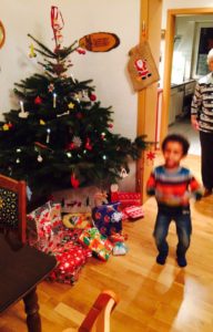 Weihnachtsbaum mit Geschenken darunter, davor ein zweijähriger Junge mit dunkler Haut